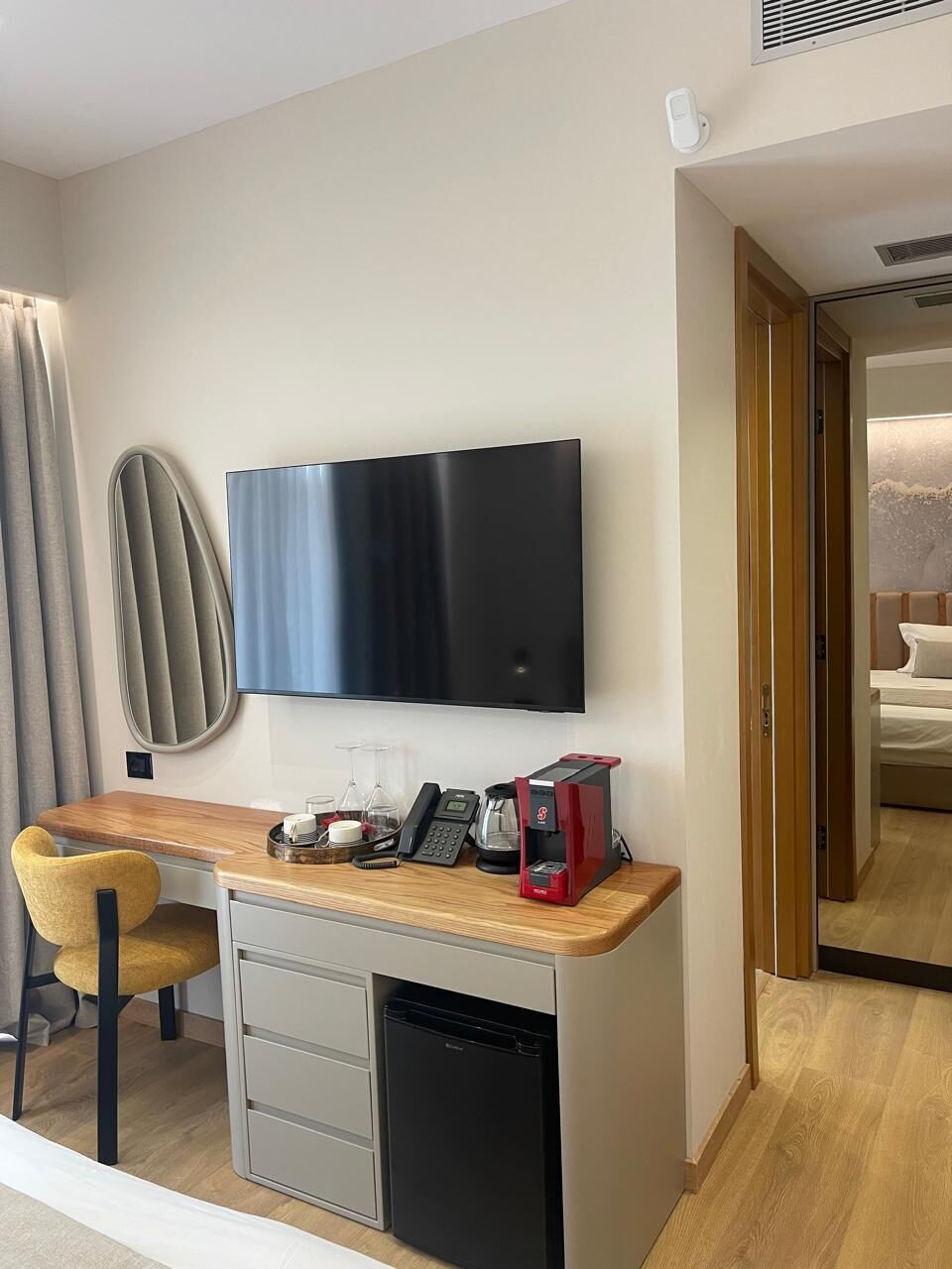 A compact hotel room with a flat-screen TV mounted on the wall, a small kitchenette with a coffee maker, a compact dishwasher, and a wooden desk with a chair.