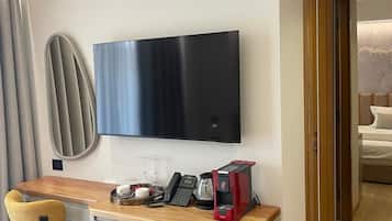 A compact hotel room with a flat-screen TV mounted on the wall, a small kitchenette with a coffee maker, a compact dishwasher, and a wooden desk with a chair.