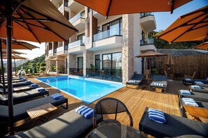 Zona de piscina con tumbonas y sombrillas, una piscina azul cristalina y un edificio moderno con balcones.