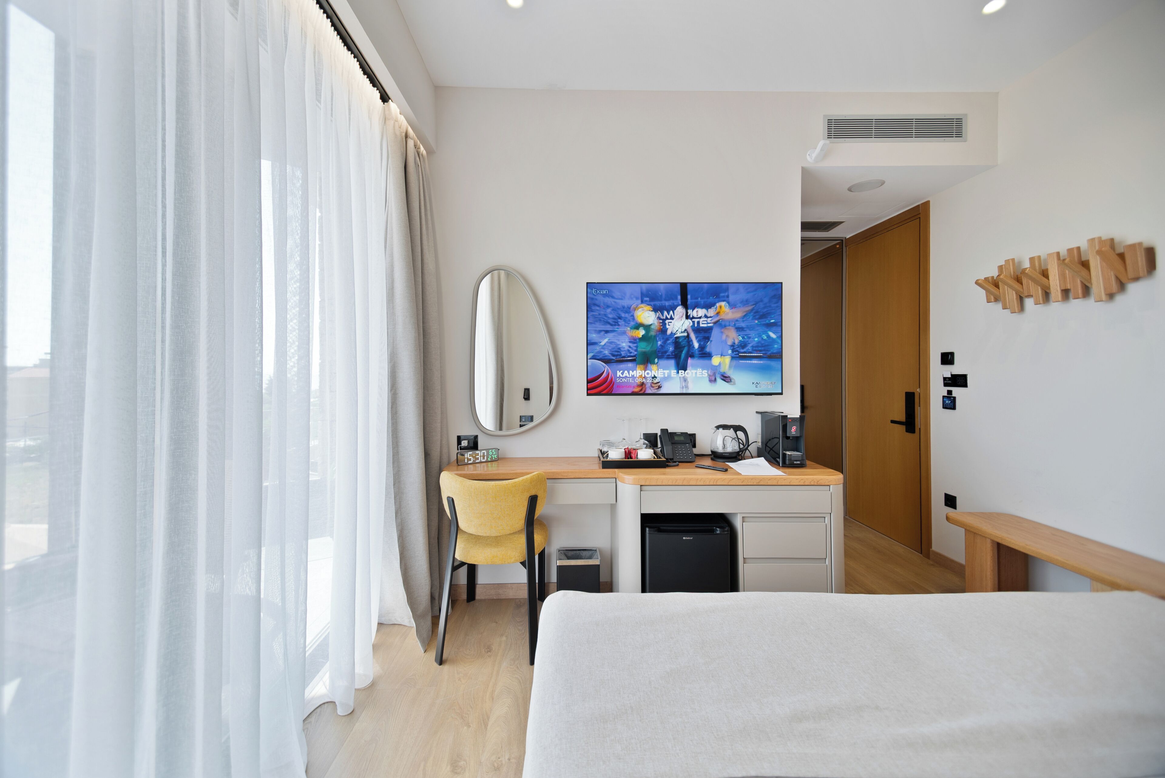 Una camera d'albergo con un letto, una scrivania con una sedia, una televisione, uno specchio e una finestra con tende.