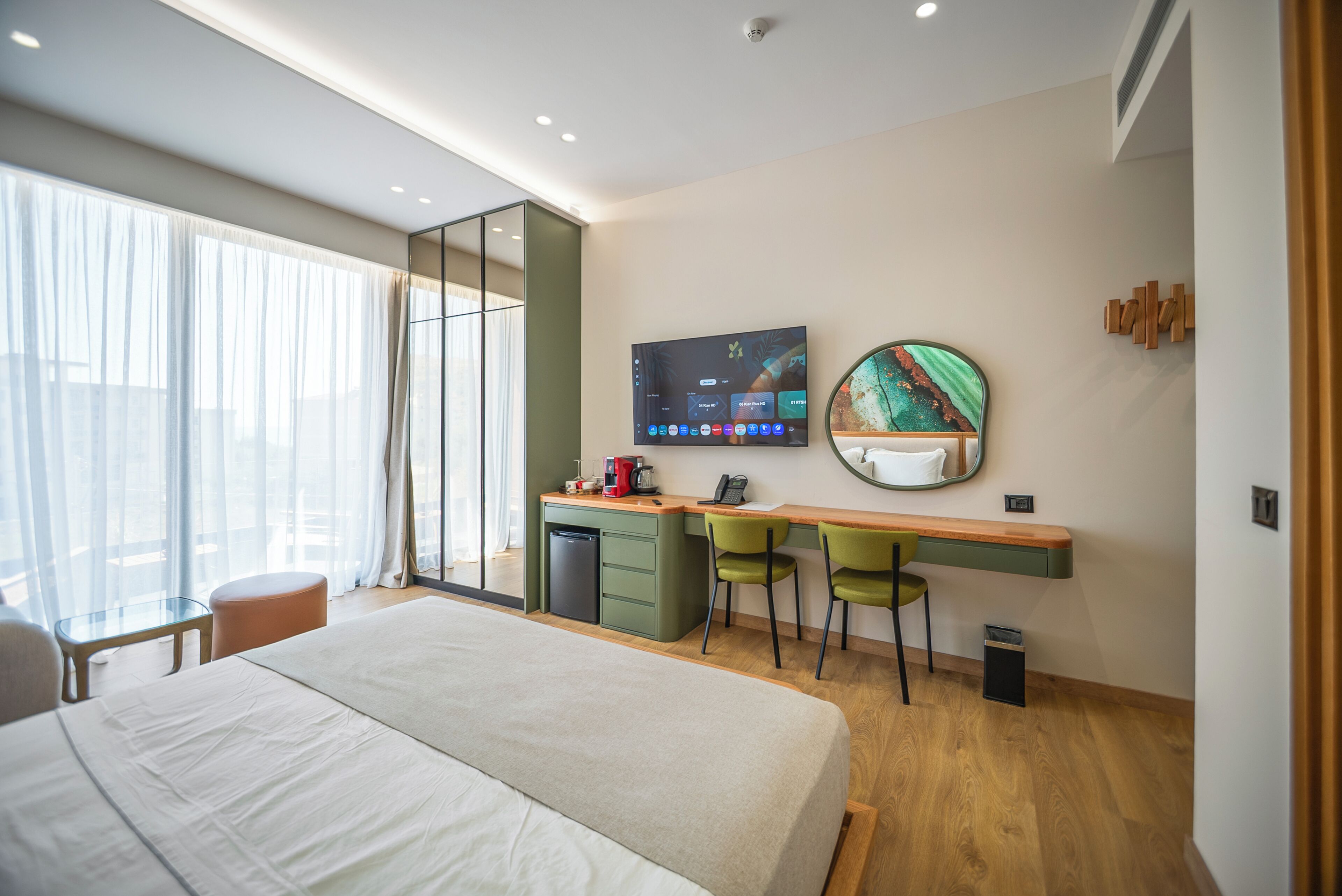 Habitación de hotel moderna con una cama grande, un escritorio con dos sillas, un televisor y un ventanal con cortinas.