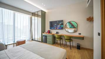 Habitación de hotel moderna con una cama grande, un escritorio con dos sillas, un televisor y un ventanal con cortinas.