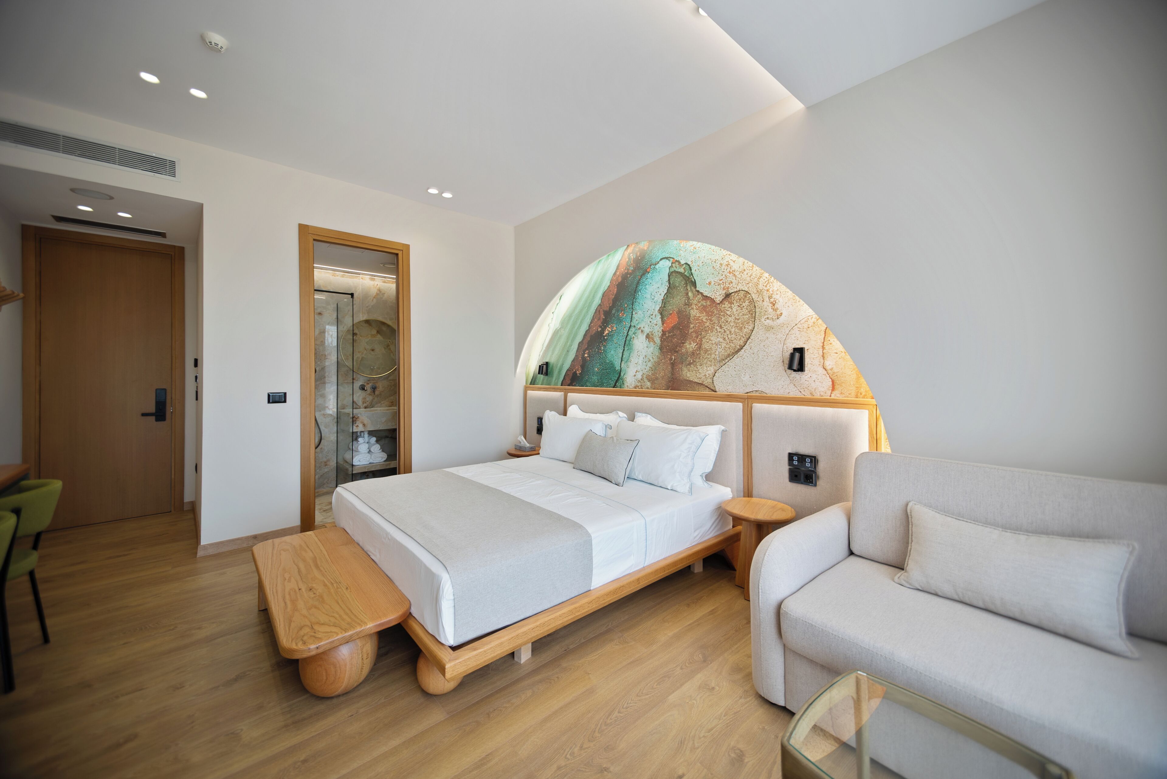 A modern hotel room with a large bed, a sofa, a wooden bench, and a glass coffee table.