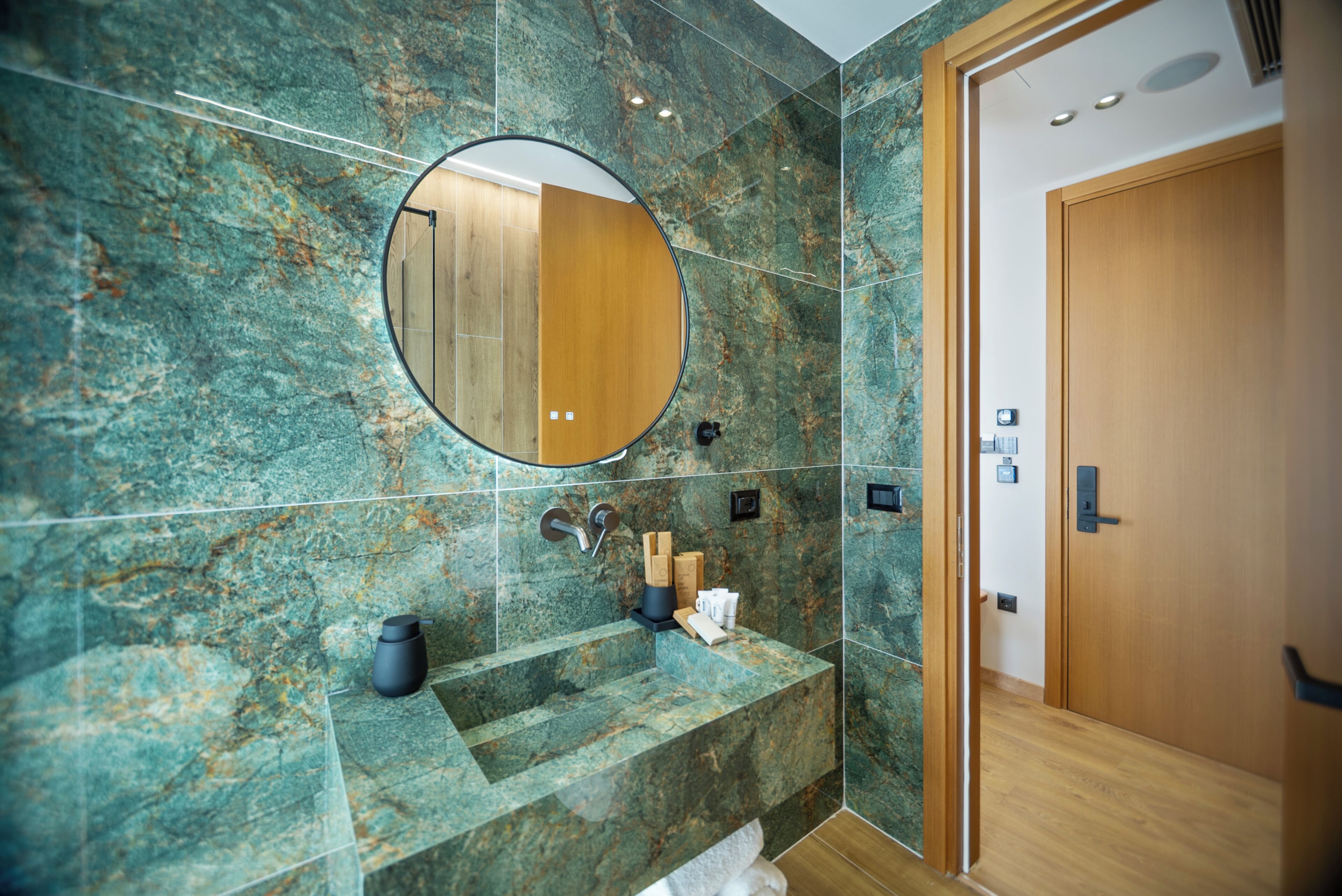 Een badkamer met een marmeren wand, een ronde spiegel, een wastafel met toiletartikelen, en een houten deur.