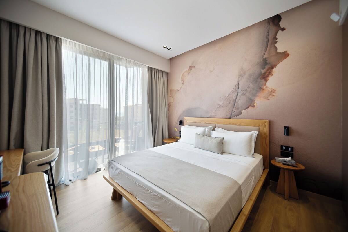A hotel room with a large bed, a desk, and a chair. There is a view of the outdoors through the window.