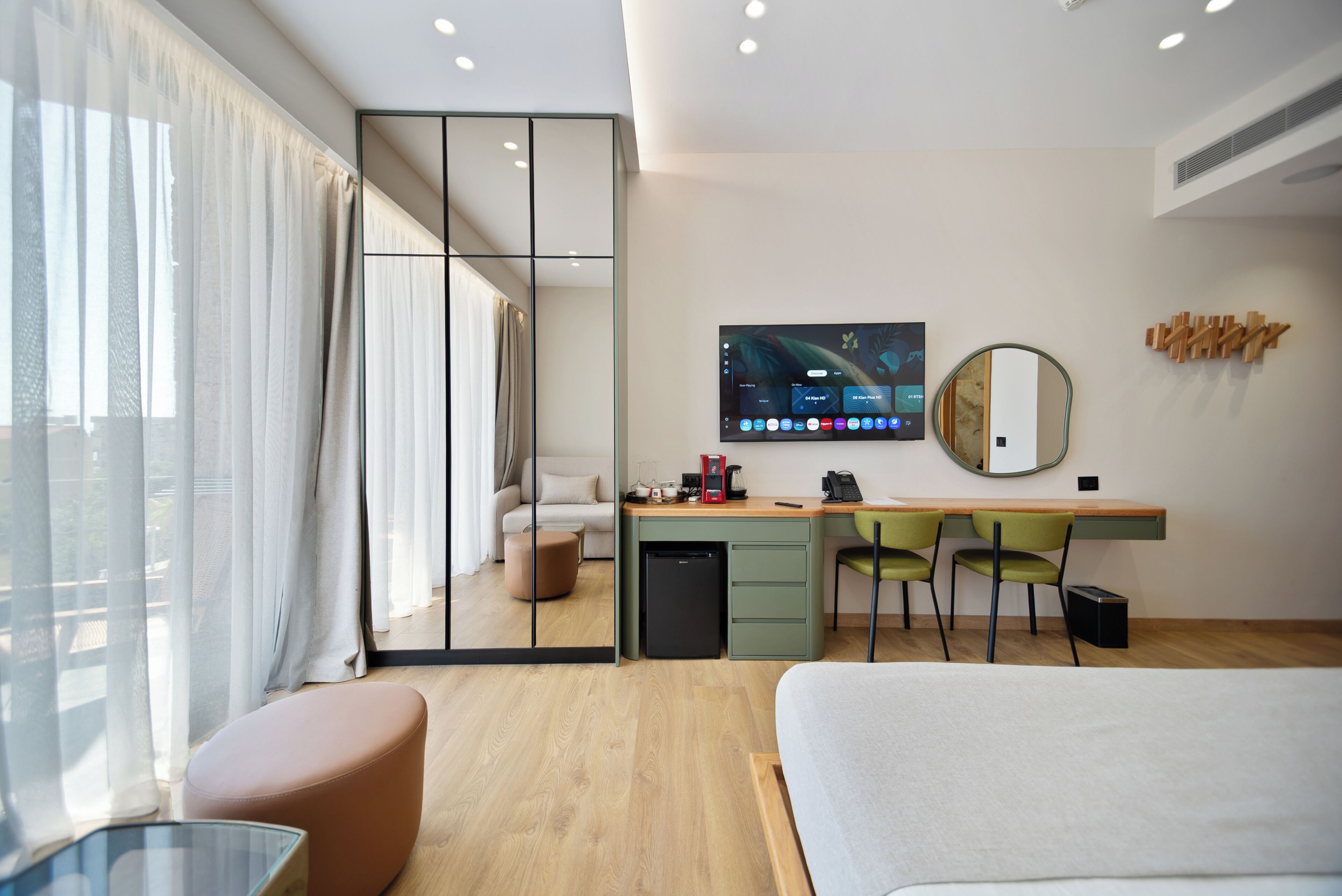 A modern hotel room with a bed, a desk with a computer, a mirror, and a view of the outside.