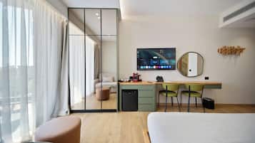 A modern hotel room with a bed, a desk with a computer, a mirror, and a view of the outside.