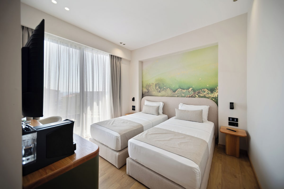 A hotel room with two beds, a TV, a painting, and a small table.