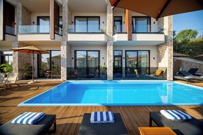 A modern two-story building with a swimming pool, lounge chairs, and umbrellas.