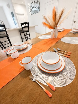 Ein Esstisch ist gedeckt mit orangefarbenen Platzsets, weißen Tellern mit orangefarbenen Rändern und passenden Tassen.
