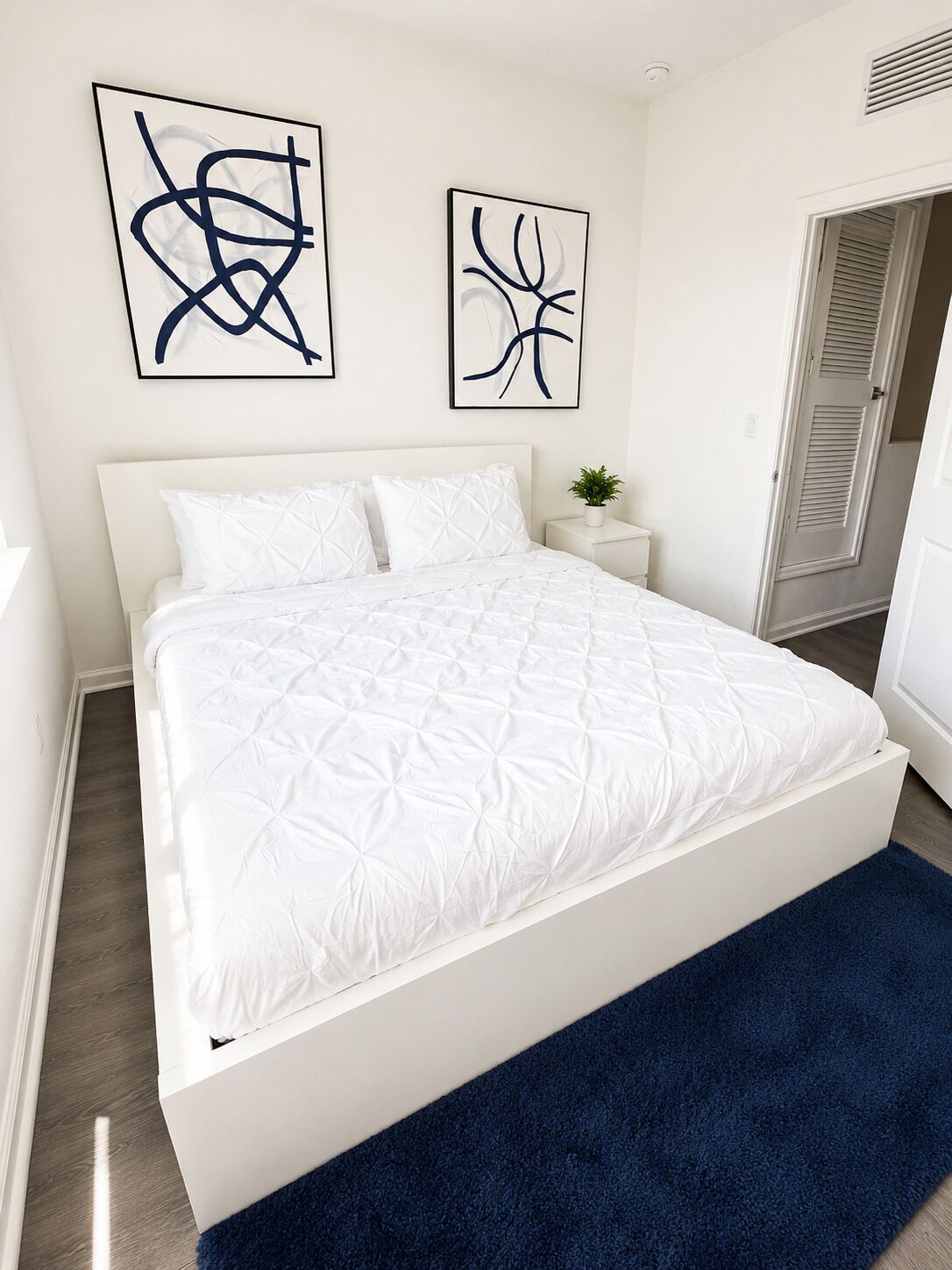 Een bed met een wit hoofdeinde en een blauw vloerkleed, twee abstracte kunstwerken aan de muur, en een klein plantje op de nachtkast.