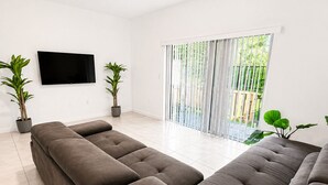 Ein modernes Wohnzimmer mit einer großen Eckcouch, einem flachbildfernseher an der Wand und einer Glastür zum Balkon.