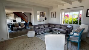 A spacious living area with a large sectional sofa, a coffee table, and a patterned floor. A staircase leads to an upper level, and there are framed pictures on the walls.
