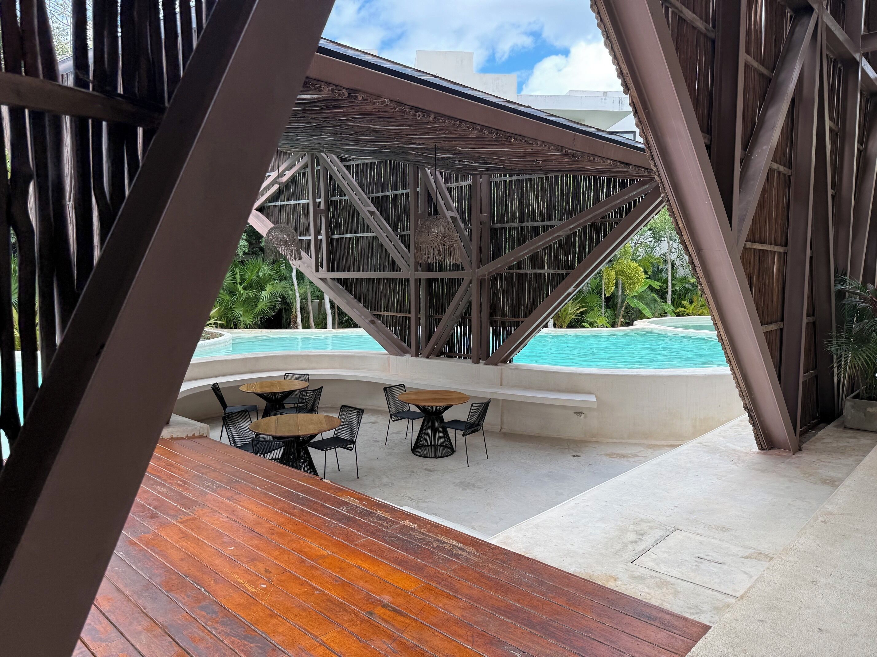 A poolside area with wooden tables and chairs, a covered seating area, and a view of a swimming pool.
