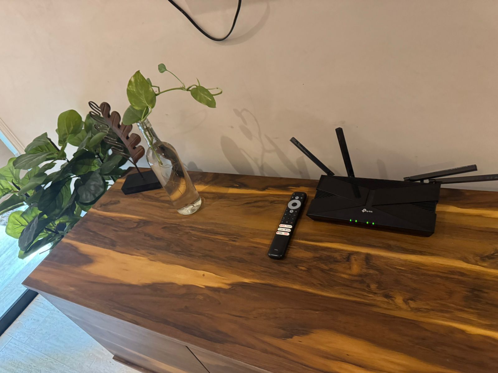 Un tavolo in legno con un vaso di vetro contenente una pianta, un telecomando e un router nero.