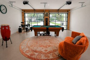 A room with a pool table, a red drum set, a clock, and a large window with a view of a house and trees. - Boho Luxury Retreat near Winter Garden & Disney - Game Room & Statement Kitchen (Ocoee)