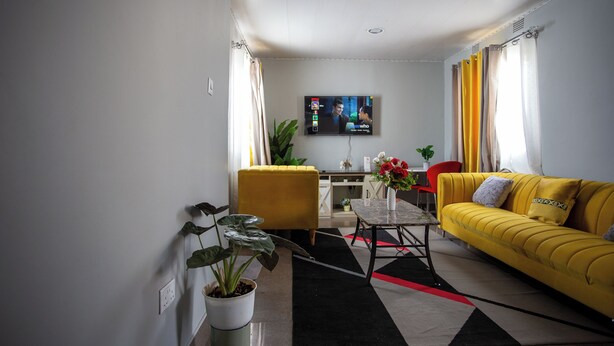 Ein Wohnzimmer mit gelber Couch, Couchtisch, an der Wand montiertem Fernseher und einer Topfpflanze.