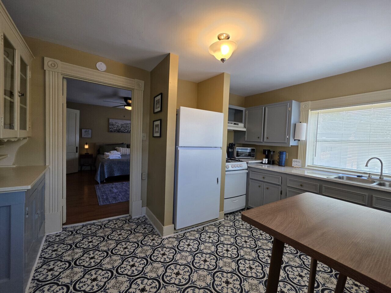 A compact kitchen with a refrigerator, microwave, stove, and sink. There is a patterned floor and a door leading to a bedroom.