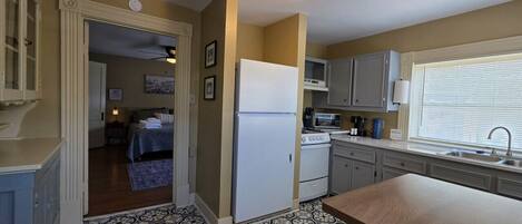 A compact kitchen with a refrigerator, microwave, stove, and sink. There is a patterned floor and a door leading to a bedroom.