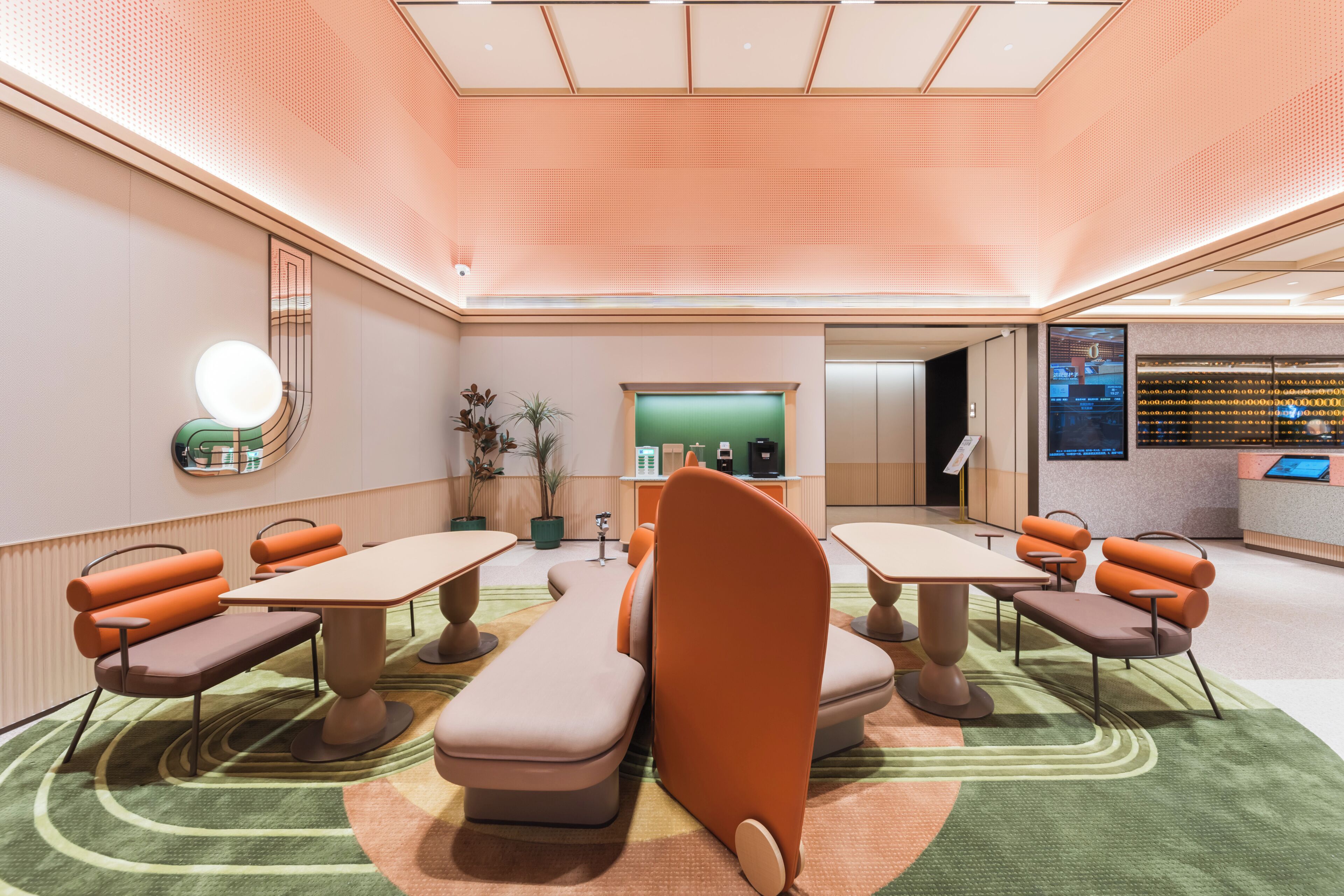 Een moderne hotellobby met een receptiebalie, een zithoek en een groene wand met een digitaal scherm.