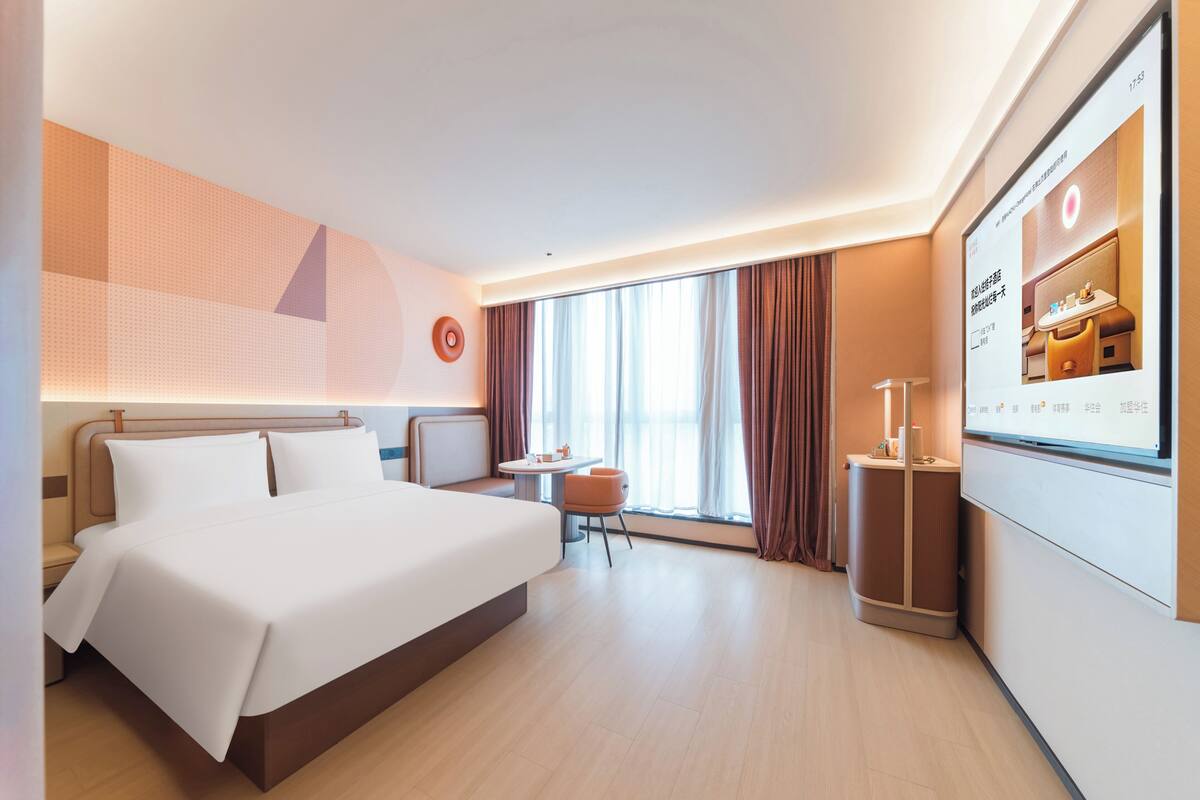 Una habitación de hotel moderna con una cama grande, una mesita, una silla y una pantalla de televisión mostrando una imagen de otra habitación.
