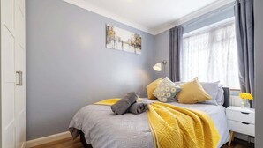 Une chambre à coucher comprenant un lit, une table de chevet, une lampe, une fenêtre avec des rideaux et une œuvre d’art murale.