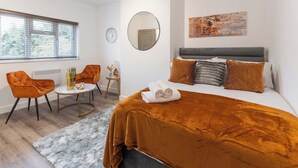 Una camera d'albergo con un letto, uno specchio rotondo, un quadro, una finestra con persiane, un tavolino con un vaso e due poltrone arancioni.