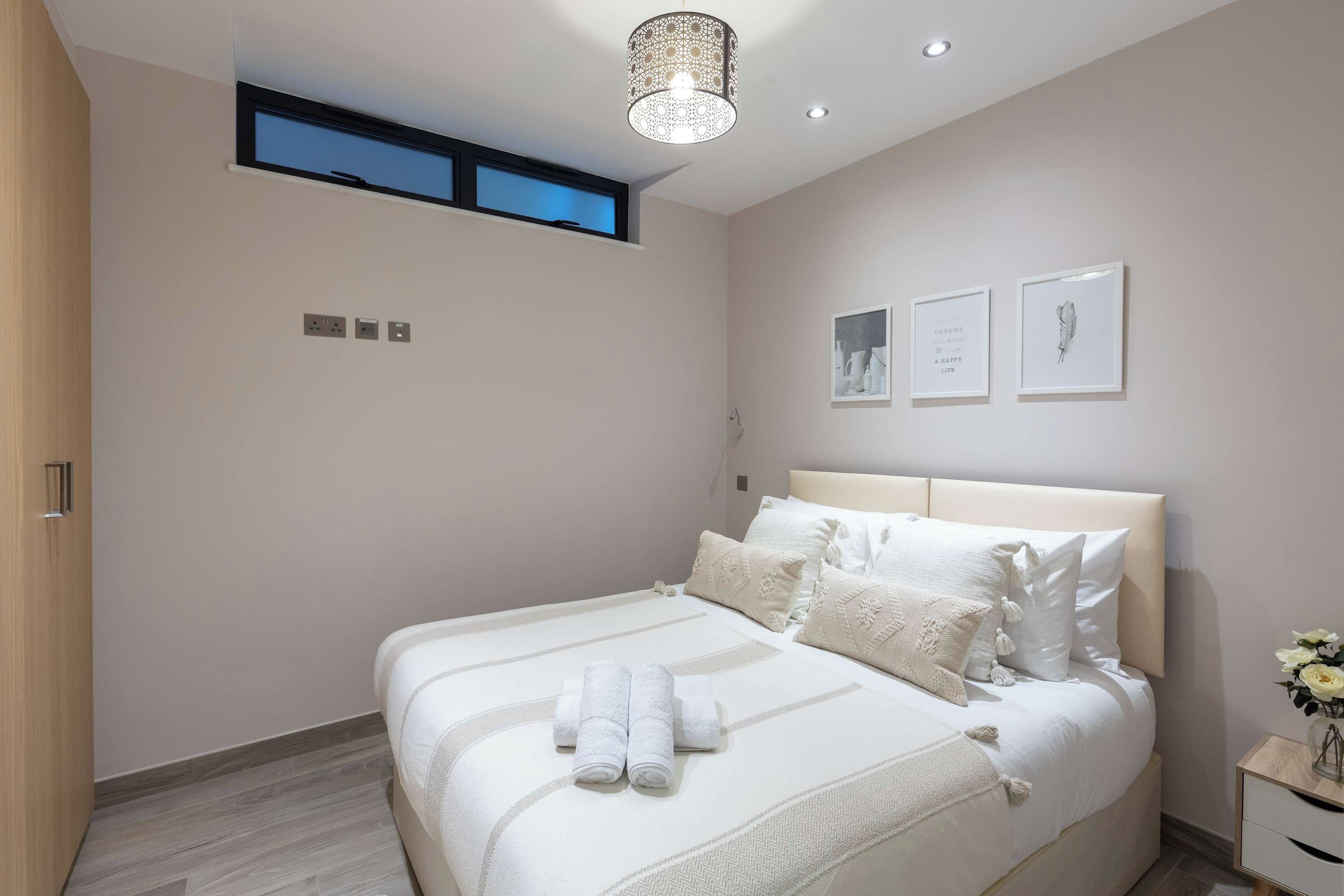 Un dormitorio con cama, mesita de noche, lámpara de pared y una ventana pequeña.