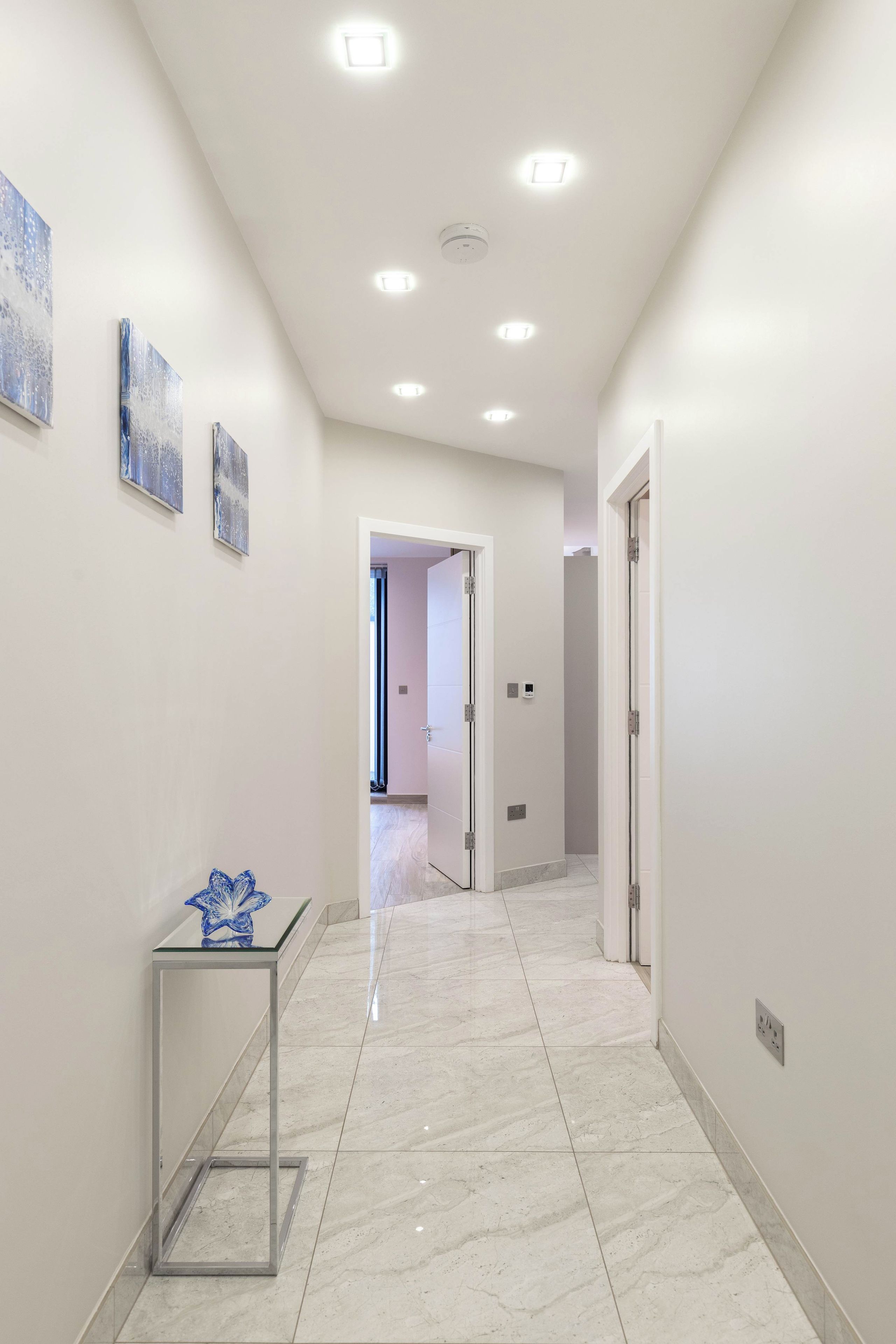 Un pasillo con paredes blancas, piso de mármol y luces empotradas en el techo. Hay puertas a ambos lados y una pequeña mesa con un jarrón azul a la izquierda.