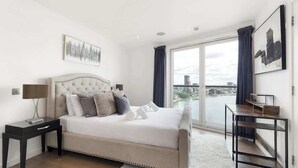 A bedroom with a large bed, a nightstand, a table, and a view of a cityscape through a large window. - Exclusive London Penthouse with River Views (Greater London)