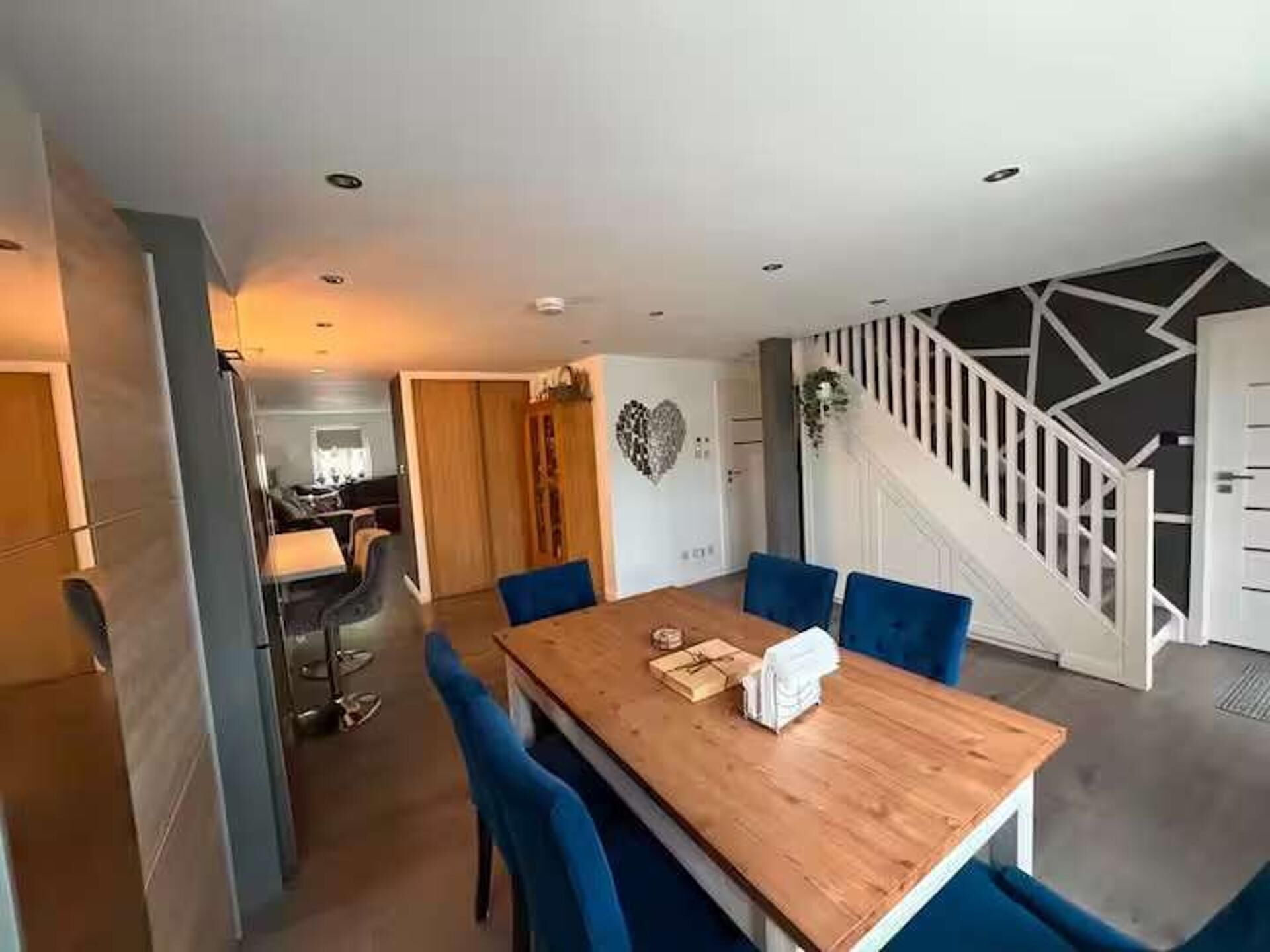 A modern dining area with a wooden table and blue chairs, a staircase with a geometric design, and a kitchen in the background.