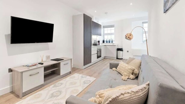 Un salon moderne avec un téléviseur à écran plat, un canapé gris, une table basse en bois et une cuisine en arrière-plan.