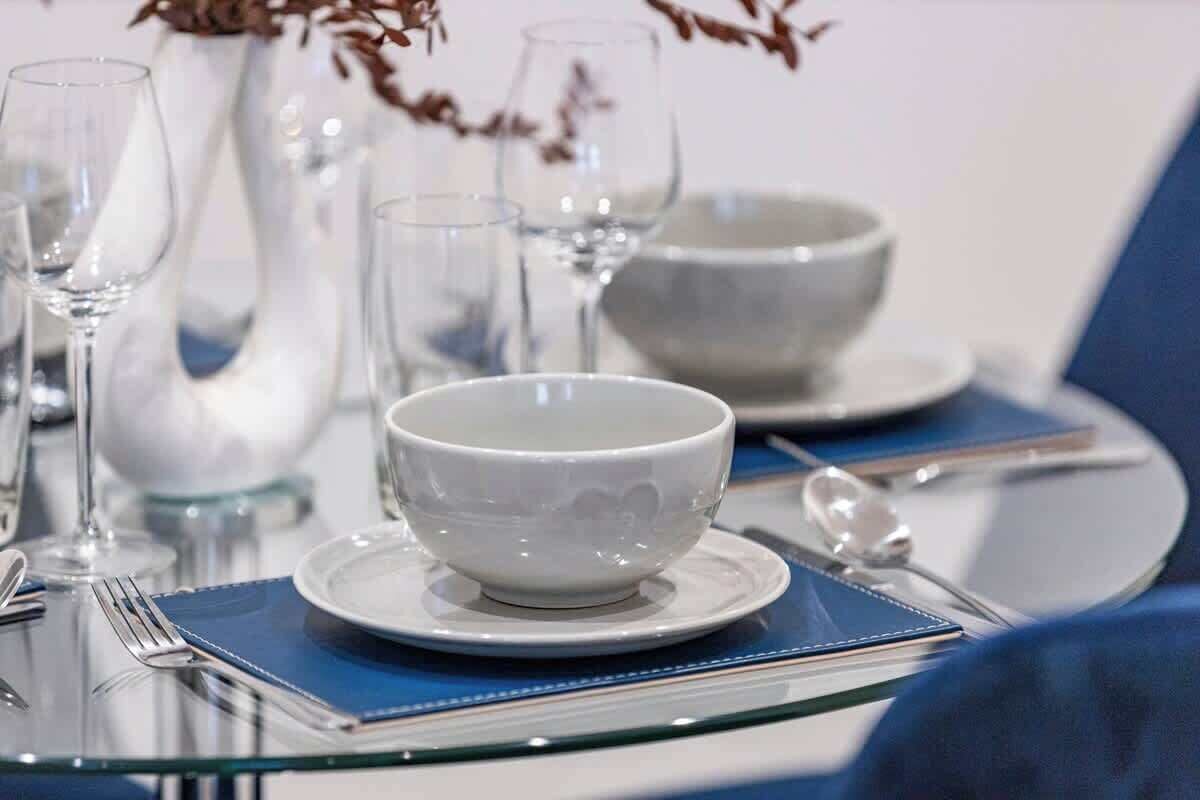 Une table à manger dressée avec des bols blancs, des verres à vin transparents et des couverts sur un chemin de table bleu.
