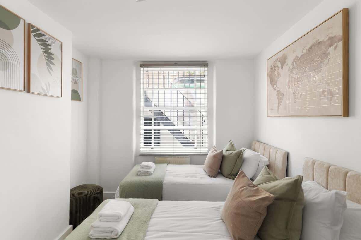 A hotel room with two beds, a window with blinds, a map on the wall, and a small stool.