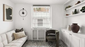 Ein modernes Wohnzimmer mit einem weißen Sofa, einem dunklen Sessel, einer Heizung, einem Fenster mit Jalousien und einem Regal mit Dekoration.
