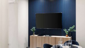 Ein modernes Zimmer mit einem an einer dunklen Wand montierten Flachbildfernseher, einem Holzkonsolentisch mit Pflanze und einem Essbereich mit Stühlen und Tisch.