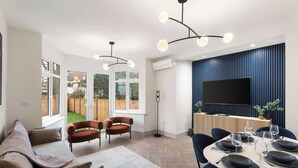 Ein modernes Wohnzimmer mit einer Couch, Sesseln, einem Esstisch für sechs Personen und einem an einer blauen Wand montierten Fernseher.