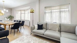 Ein modernes Wohnzimmer mit einer Couch, einem Esstisch, Stühlen, großen Fenstern mit Vorhängen und einer Stehlampe.