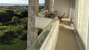 Ein Balkon mit Glasgeländer, einem Esstisch mit Stühlen und Blick auf das Meer und Grünflächen.