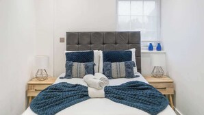 Ein ordentlich bezogenes Bett mit grauem Kopfteil, zwei Nachttische mit Lampen und ein Fenster mit Jalousien.