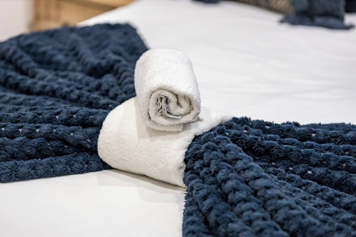 A bed with a white sheet, a rolled white towel, and a dark blue textured blanket.