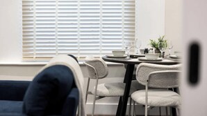 Ein Essbereich mit Tisch für zwei Personen, Stühlen, einem Fenster mit Jalousien und einer Couch im Vordergrund.