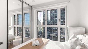 A modern hotel room with a large bed, a wooden bedside table with towels, a large mirror, and a view of high-rise buildings through floor-to-ceiling windows. - Stylish Central London Flat Steps from the Tube (Greater London)