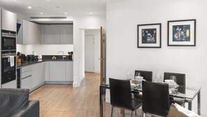 A modern kitchen with grey cabinets, a dining area with a glass table and black chairs, and two framed pictures on the wall. - Stylish Central London Flat Steps from the Tube (Greater London)