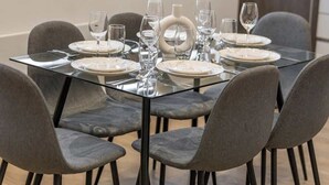 Una mesa de comedor dispuesta con platos, vasos y cubiertos, rodeada de sillas tapizadas en gris.