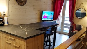 A kitchen with a wooden island, a TV displaying a news program, and a dining area with red curtains and a mirror.