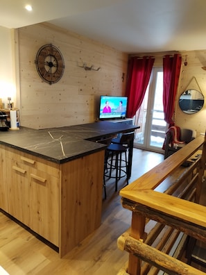Een keuken met een houten eiland, een televisie waarop een nieuwsuitzending te zien is, en een eethoek met rode gordijnen en een spiegel.