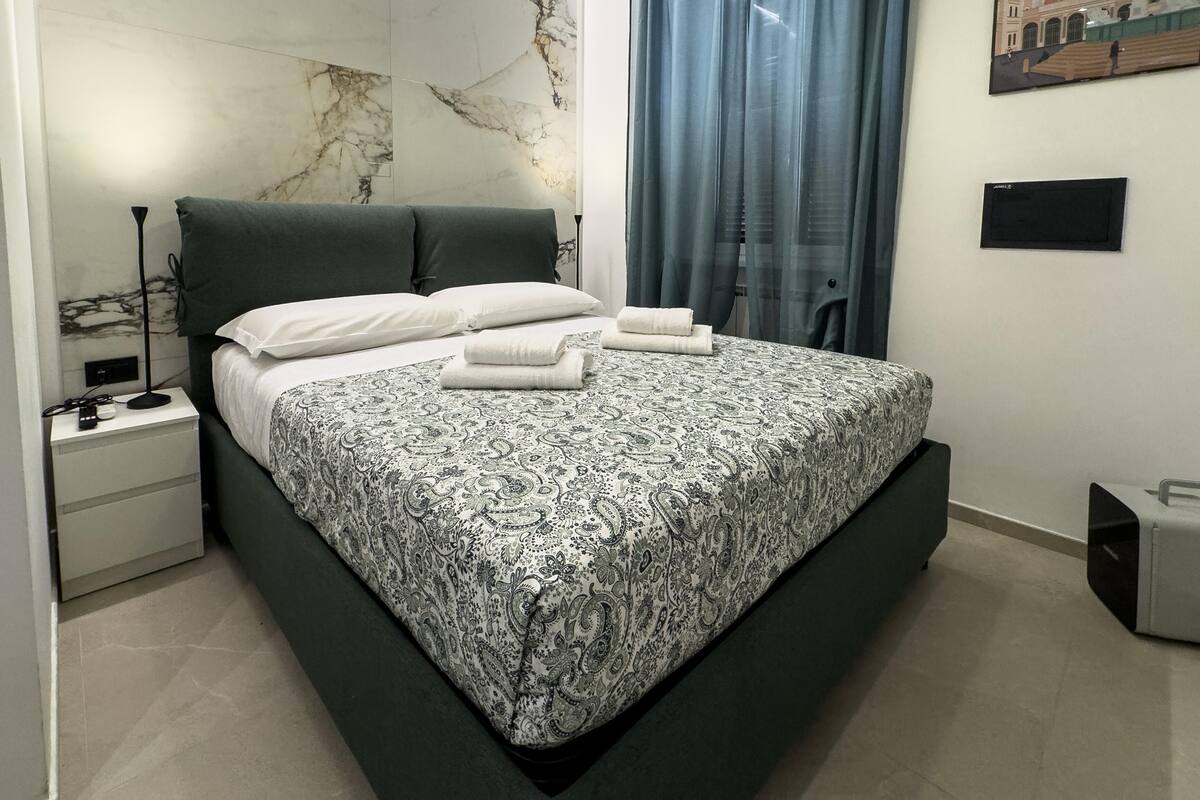 Un dormitorio moderno con cabecera de mármol, una cama con ropa de cama estampada, una mesita de noche, una lámpara, una ventana con cortinas y una obra de arte colgada en la pared.