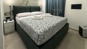 A modern bedroom with a marble headboard, a bed with patterned bedding, a nightstand, a lamp, a window with curtains, and a wall-mounted artwork.