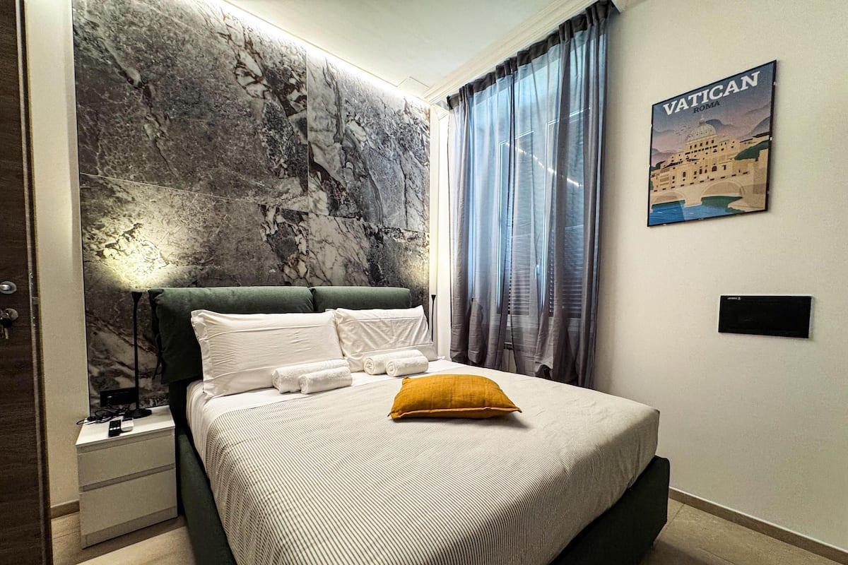 Un dormitorio con cama, mesita de noche, lámpara, pared con textura similar al mármol y un cuadro enmarcado de Roma, Ciudad del Vaticano.